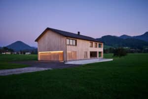 Zweigeschossiges Einfamilienhaus mit Holzfassade und großflächigen Fenstern inmitten einer grünen Wiese, Abendstimmung mit Bergkulisse.