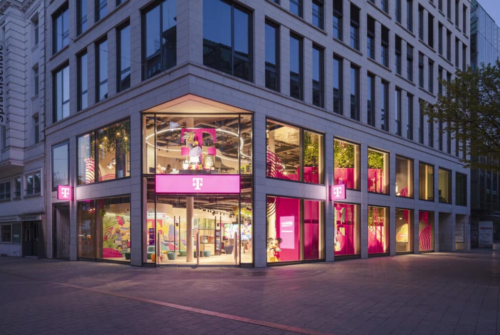 Außenansicht des modernen Telekom-Flagshipstores bei Abendlicht mit großen Glasfassaden und beleuchtetem Magenta-Logo.