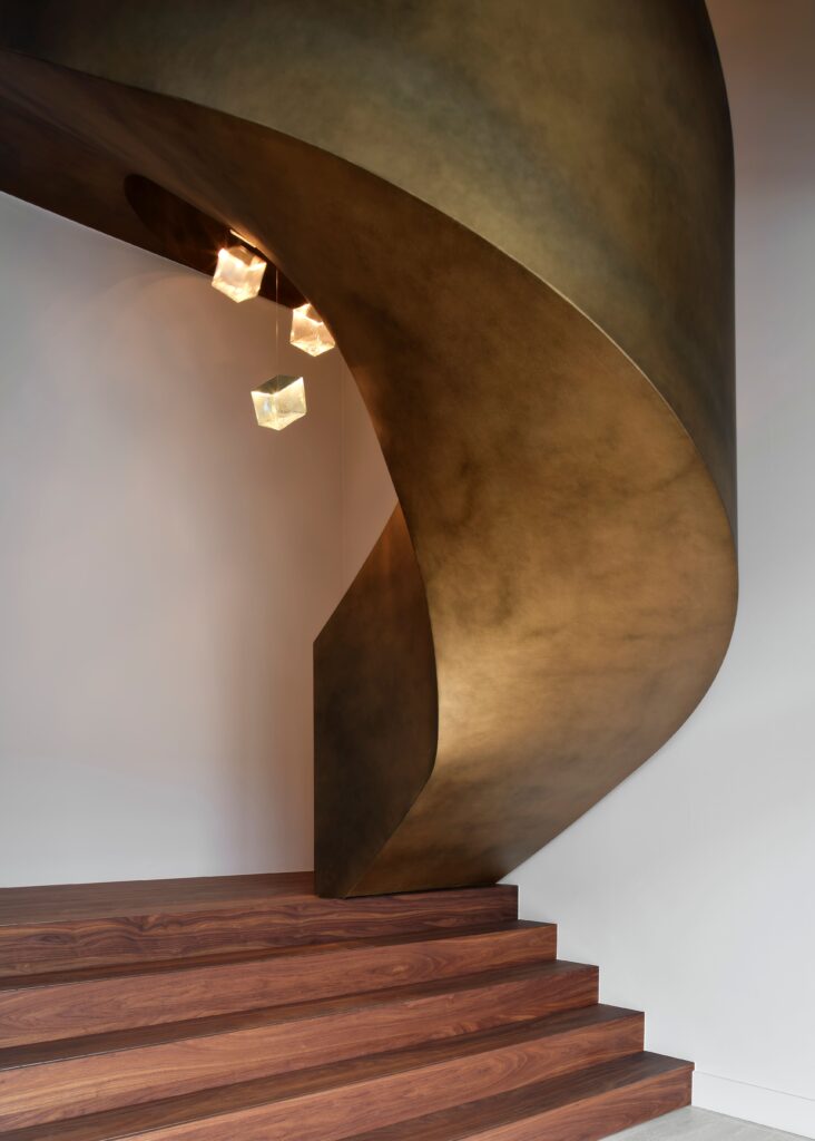 Elegante, skulpturale Wendeltreppe mit bronzefarbener Oberfläche und massiven Holzstufen, ergänzt durch moderne würfelförmige Pendelleuchten.