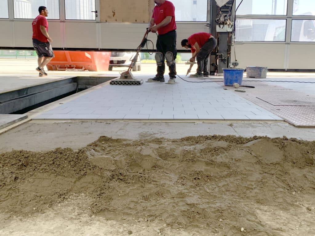 Handwerker verlegen einen Industrieboden im Rüttelverfahren, tragen Dickbettmörtel auf und verdichten die frisch verlegten Fliesen mit einer Rüttelmaschine.