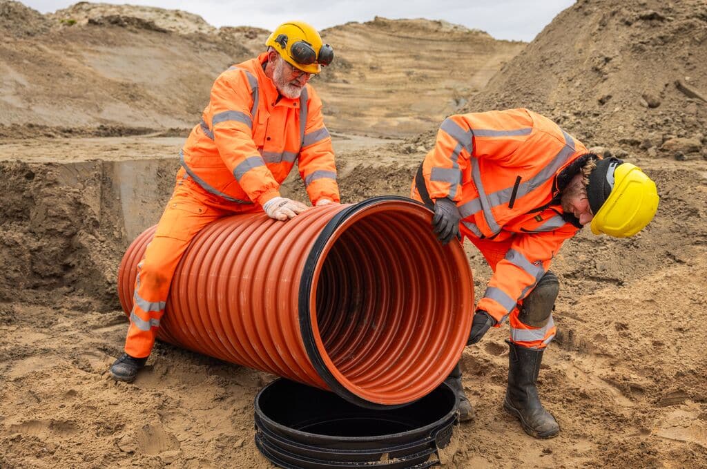 Zwei Bauarbeiter in Warnschutzkleidung installieren ein großes orangefarbenes Wavin Tegra 600 Schachtrohr in einer Sandbaugrube.