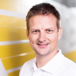 Portraitaufnahme von Jonas Steigmiller, Ingenieur bei best wood Schneider, mit weißem Poloshirt vor gelb-weißer Wand.