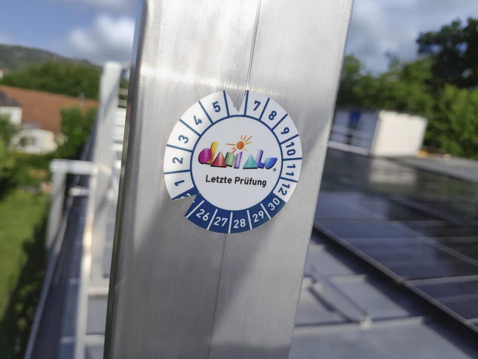 Nahaufnahme einer Prüfplakette auf einem Geländer zur Dokumentation der letzten Wartung einer Absturzsicherung