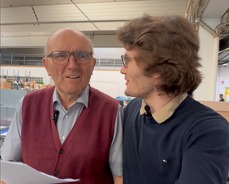 Zwei Männer stehen nebeneinander in einer hellen Industriehalle. Der ältere Mann lächelt in die Kamera, der jüngere schaut ihn an.