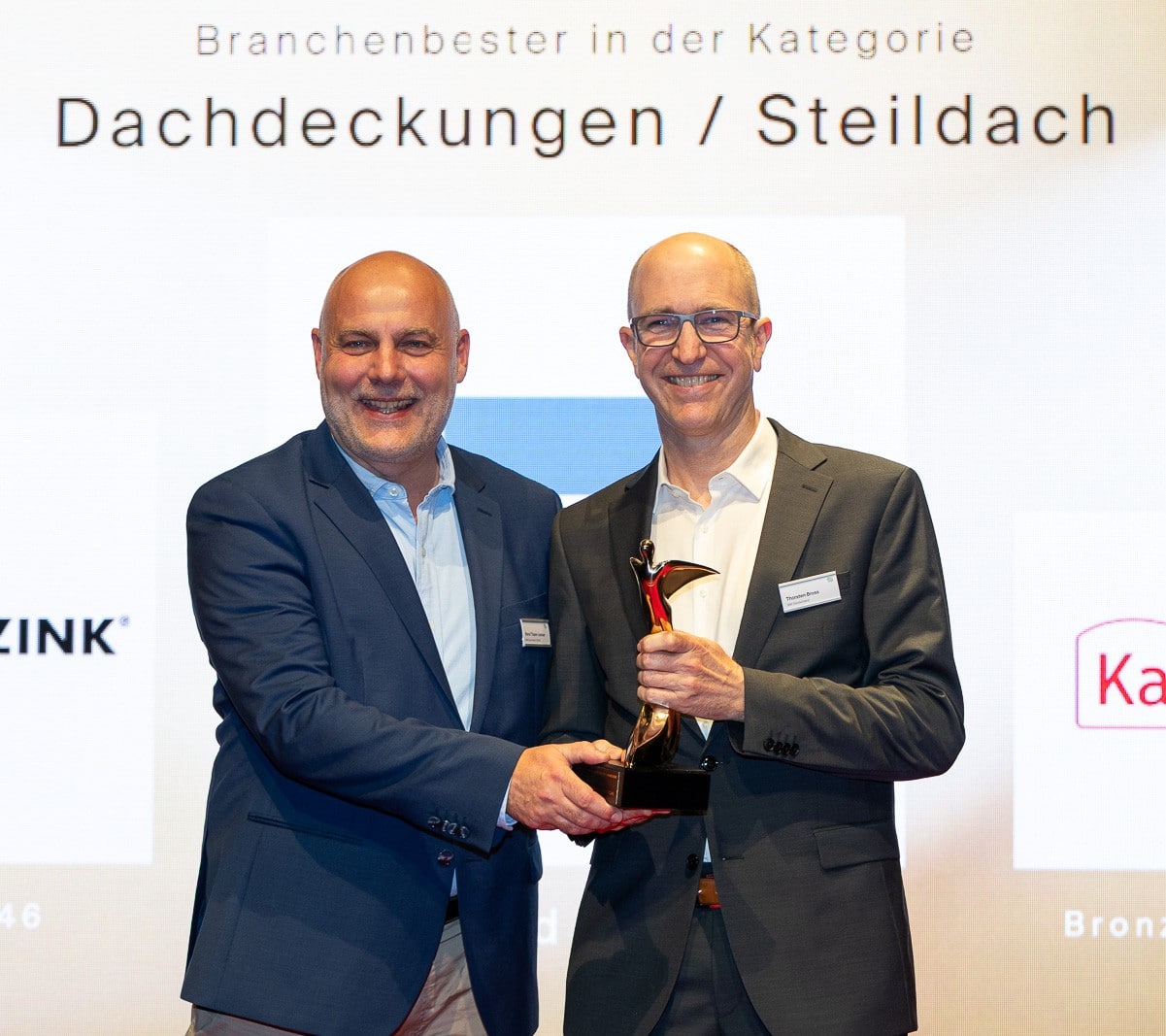 Zwei Männer auf einer Bühne halten gemeinsam eine Trophäe, die für den Branchensieg in der Kategorie Dachdeckungen / Steildach überreicht wird.
