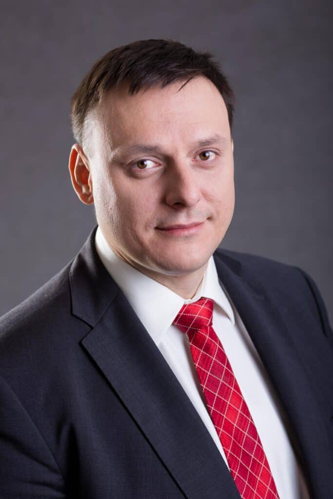 Porträt von Jiří Malkus im dunklen Anzug mit weißem Hemd und roter Krawatte vor neutralem grauen Hintergrund