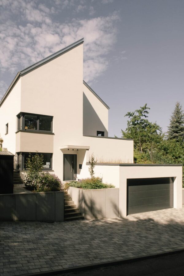 Zeitgenössisches Einfamilienhaus mit heller Putzfassade, dunklen Fensterrahmen und vorgelagerter Garage, umgeben von bepflanzten Terrassen und Treppen.