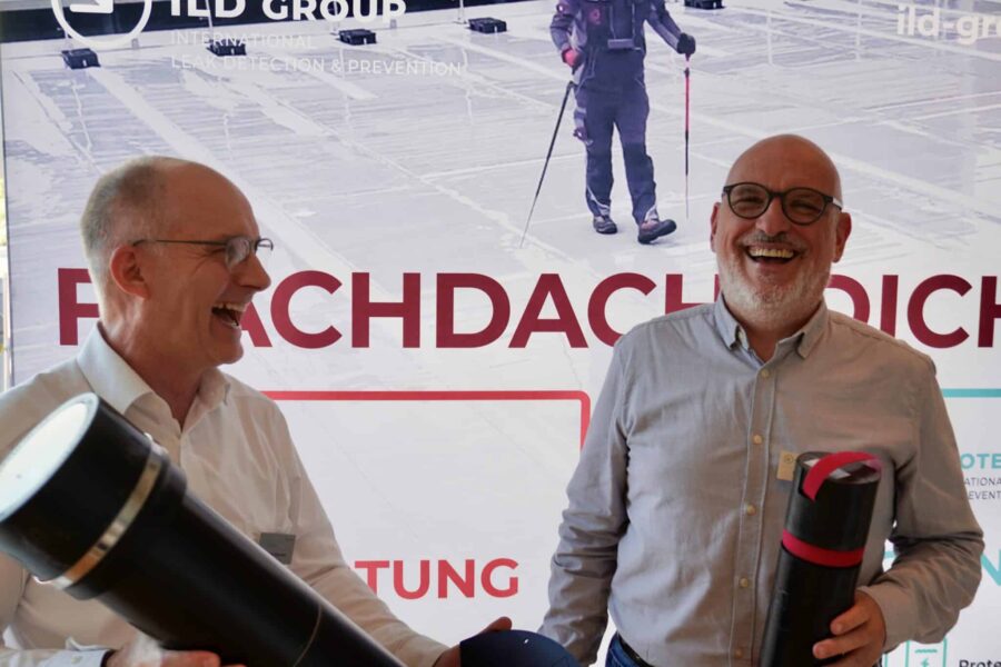 Zwei lachende Geschäftsführer von ProtectSys stehen vor einem Messebanner zur Flachdachabdichtung und halten Prüf- bzw. Messgeräte in der Hand.