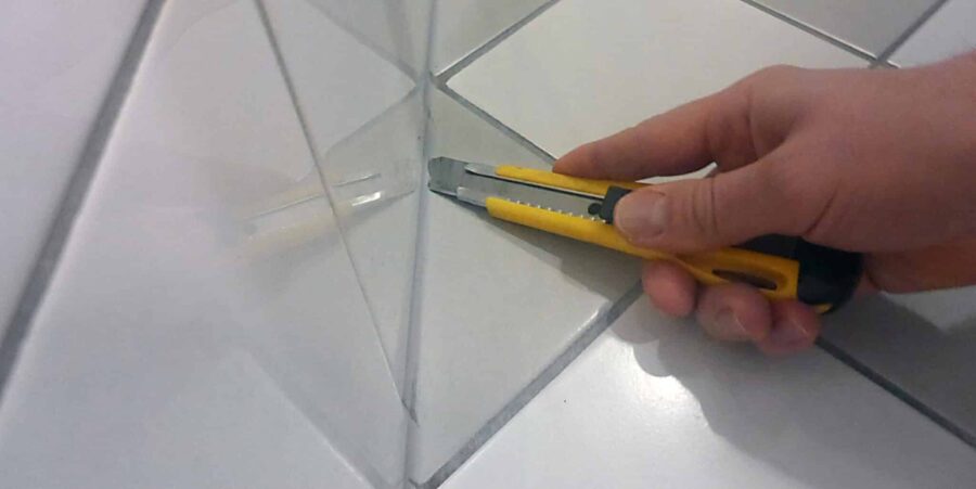 Hand führt ein Cuttermesser entlang einer Silikonfuge zwischen Wand- und Bodenfliesen, um die Fuge fachgerecht herauszuschneiden.