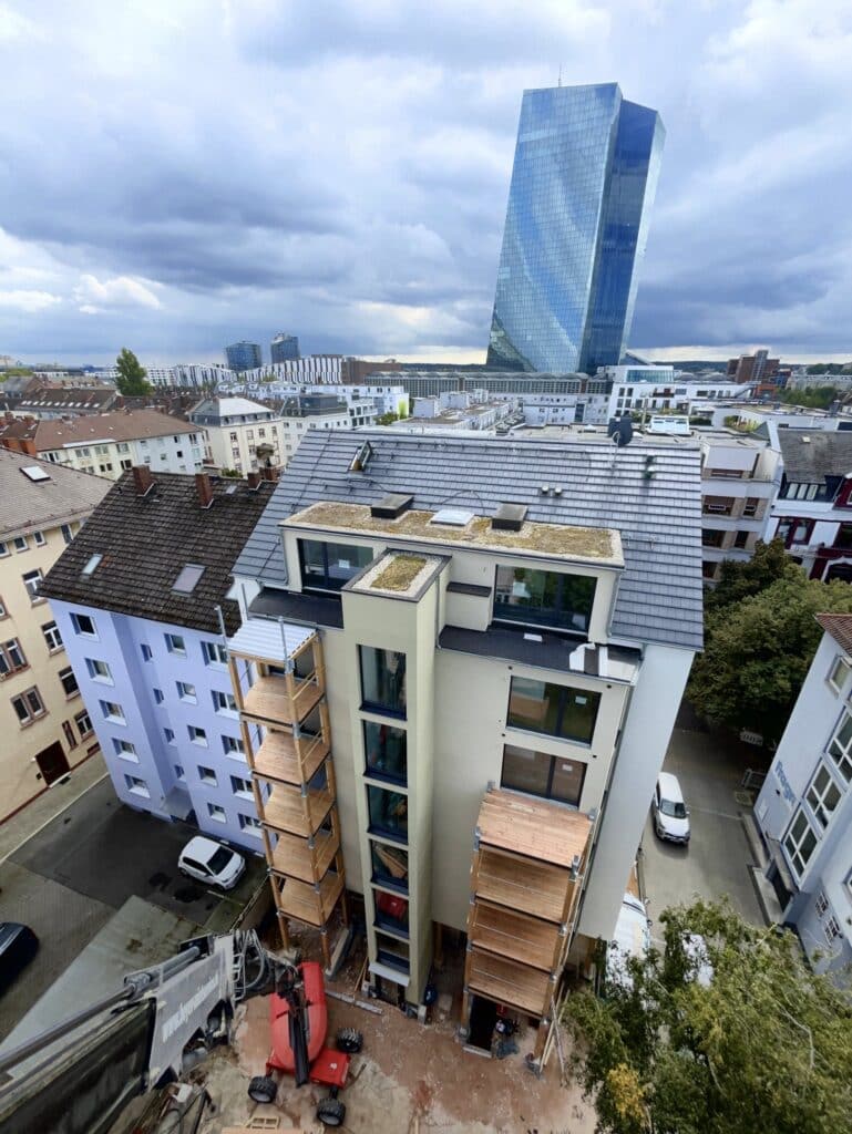 Mehrgeschossige Wohnhaus-Aufstockung im dichten Stadtgebiet von Frankfurt, im Hintergrund der gläserne EZB-Turm unter wolkenverhangenem Himmel.