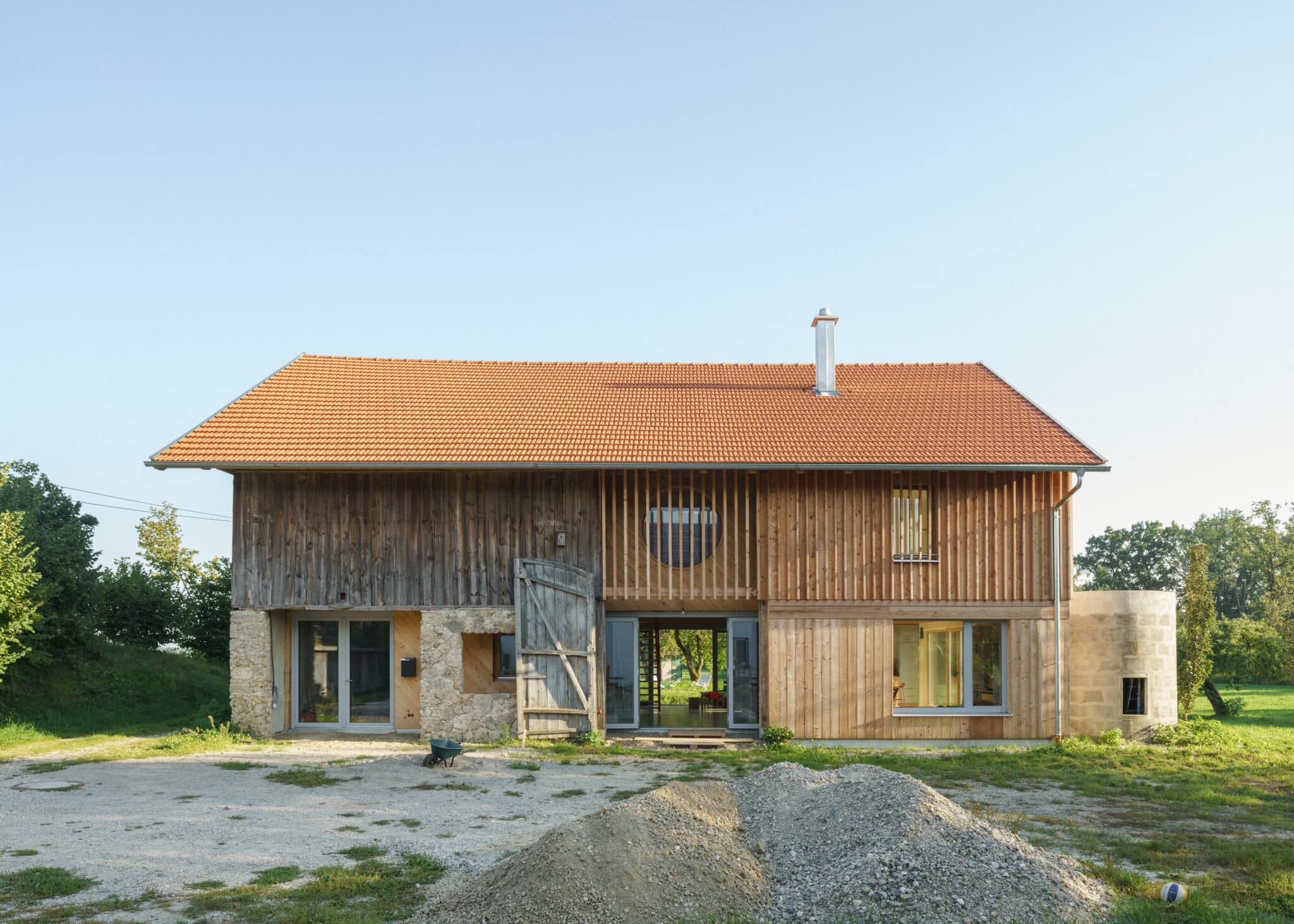 Außenansicht eines renovierten Bauernhauses mit Holzfassade, Natursteinmauerwerk und großer zentraler Öffnung zum Garten.