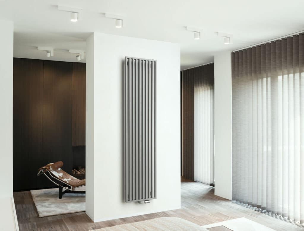 Ein schlanker, elektrisch betriebener Design-Heizkörper in Beige hängt in einem hellen, modernen Badezimmer mit Holzfußboden und großen Fensterjalousien.