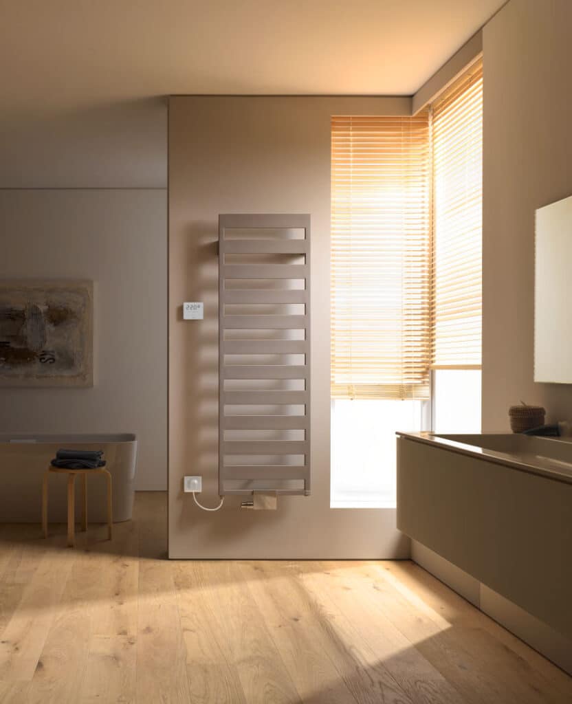 Ein hoher, vertikaler Design-Heizkörper in Grauton ist an einer weißen Wand im Wohnbereich montiert, umgeben von modernem Interieur und bodentiefen Vorhängen.