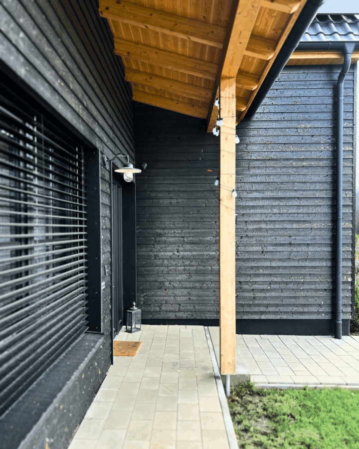 Detailaufnahme des überdachten Eingangsbereichs mit Holzstützen, dunkler Holzfassade und gepflastertem Boden.