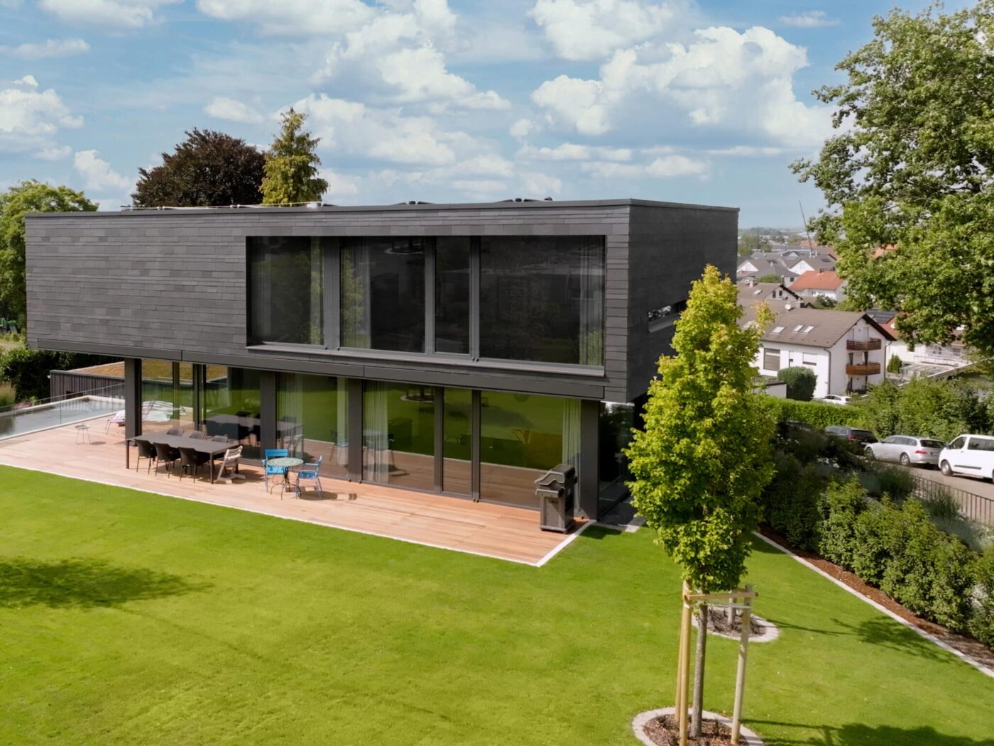 Zweigeschossiges modernes Wohnhaus mit dunkler Fassadenverkleidung, großer Holzterrasse und bodentiefen Fenstern; weitläufiger, gepflegter Garten im Vordergrund.