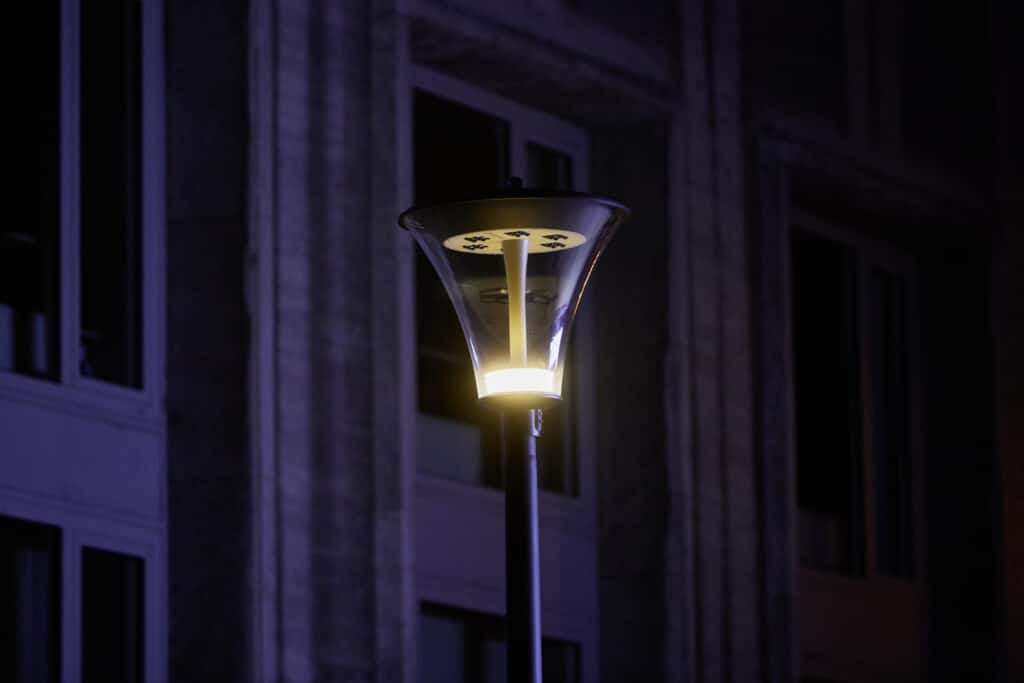 Moderne Straßenleuchte mit transparentem konischem Leuchtenkopf vor einer Fassadenfront, nachts leuchtend in warmem Licht.