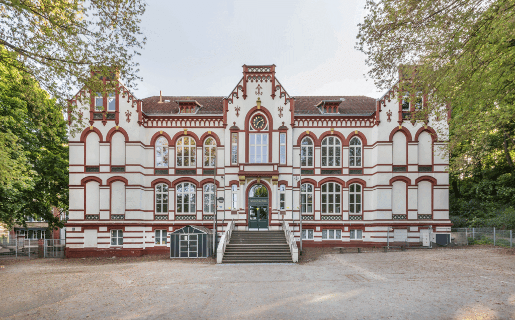 Frontansicht eines historischen Schulgebäudes mit rotem Ziermauerwerk, großen Rundbogenfenstern und zentralem Eingang, umgeben von Bäumen und einem Vorplatz.