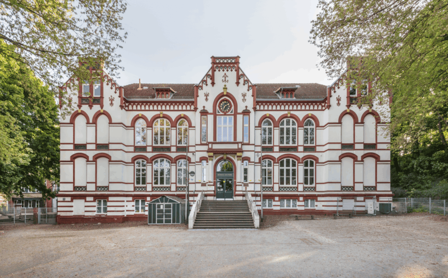 Frontansicht eines historischen Schulgebäudes mit rotem Ziermauerwerk, großen Rundbogenfenstern und zentralem Eingang, umgeben von Bäumen und einem Vorplatz.