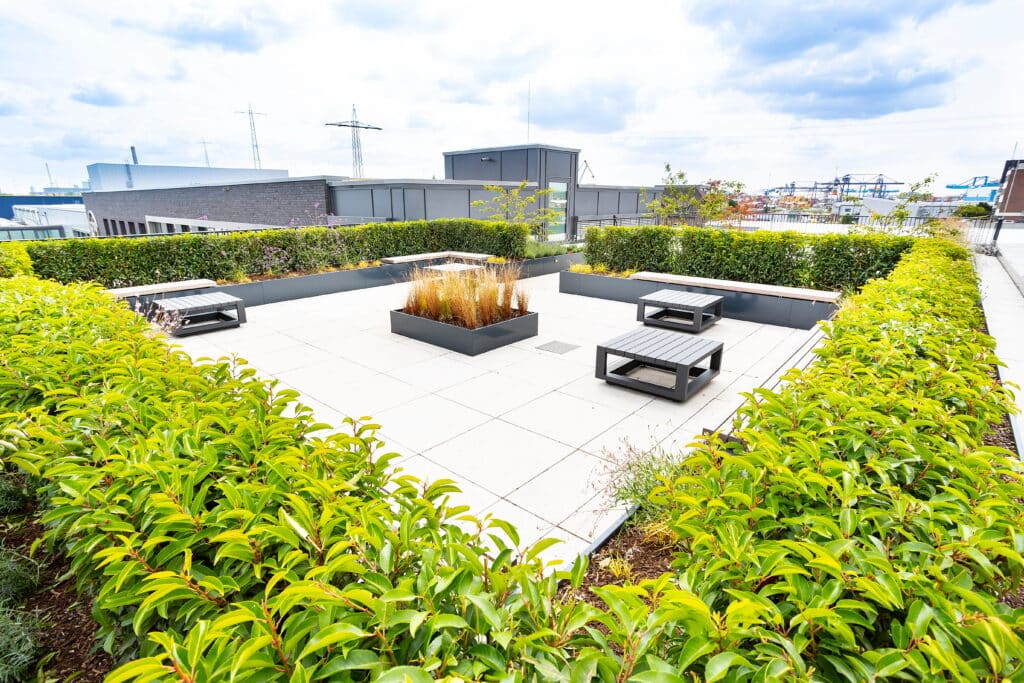 Blick auf einen rechteckigen Dachgarten mit üppigen Hecken, zentralem Pflanzbeet und mehreren grauen Sitzgelegenheiten