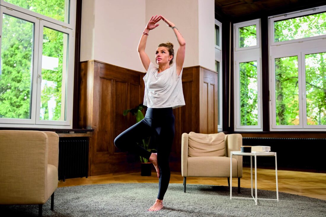 Frau macht Yogaübungen im Wohnzimmer mit stilvollem Zehnder Charleston Heizkörper vor großen Fenstern.
