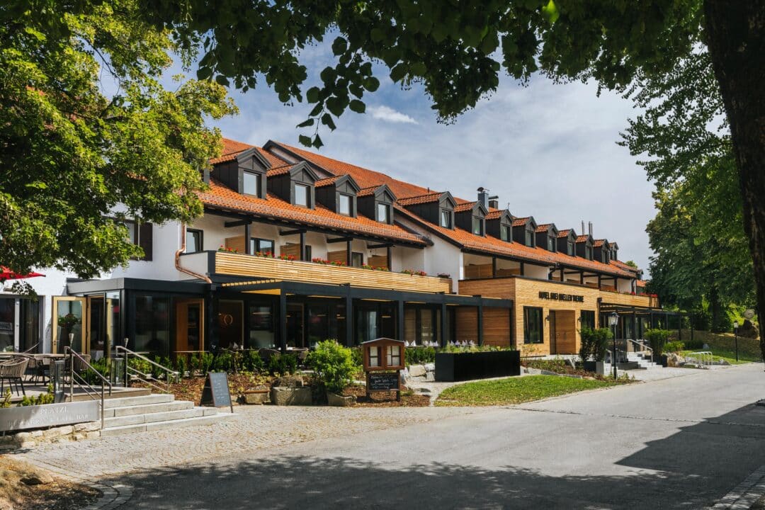 Außenansicht eines Hotels mit Terrasse, Restaurantbereich und begrüntem Umfeld.