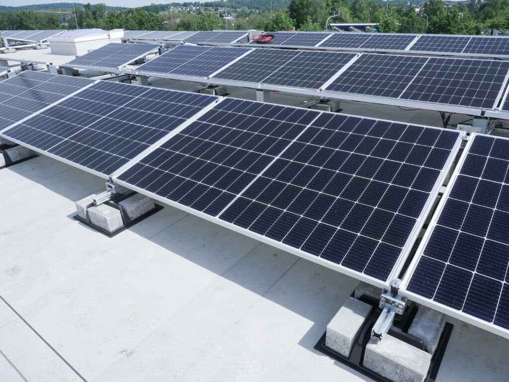 Auf einem Flachdach installierte Photovoltaik-Module mit Aufständerung und Ballastierung, im Hintergrund Bäume und Gebäudestrukturen.