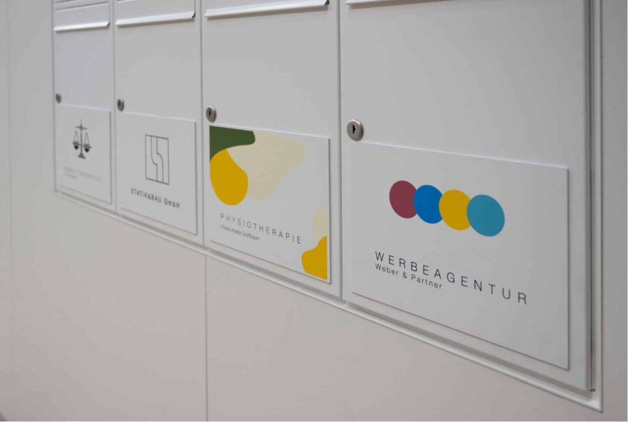 Mehrere weiße Firmenschilder in einem Innenbereich, darunter Logos einer Anwaltskanzlei, eines Statikbüros, einer Physiotherapiepraxis und einer Werbeagentur, sauber in eine Wandbeschilderung integriert.