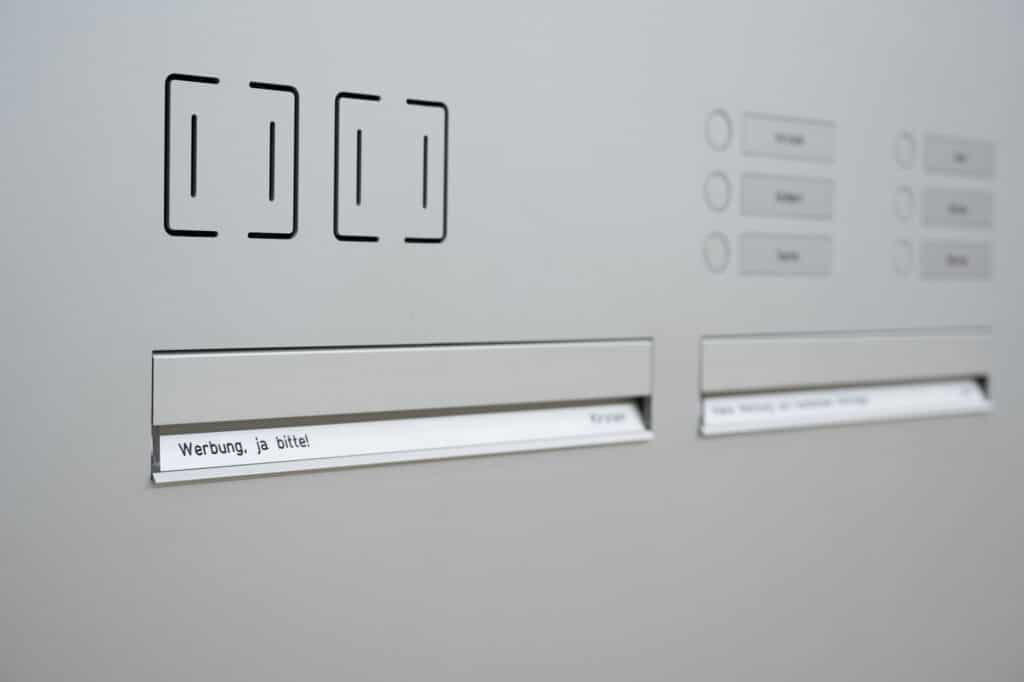 Hellgraue flächenbündige Briefkastenanlage mit integrierten Beschriftungsfeldern, darunter ein Briefschlitz mit der Aufschrift „Werbung, ja bitte!“ und weiteren Namensfeldern.