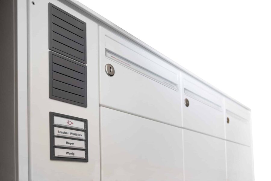 Weiße Briefkastenanlage mit grauen Lüftungs- oder Lautsprechermodulen und mehreren Namensschildern, darunter auch Klingeltasten mit Beschriftung.