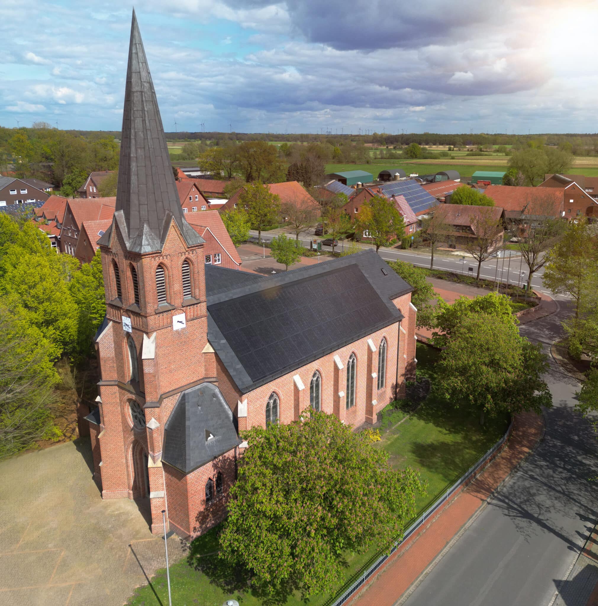 Luftaufnahme einer Backsteinkirche mit großflächig integrierter Solardachanlage inmitten eines Dorfes.