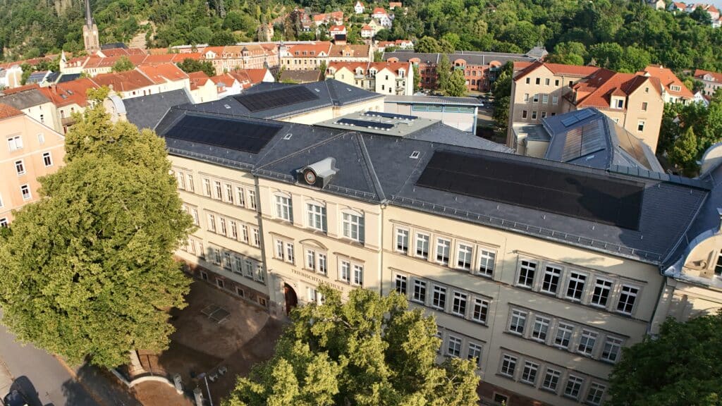 Historisches Schulgebäude in Meißen mit modernem Schieferdach und integrierter Solartechnik, umgeben von Altstadt und Grünflächen.