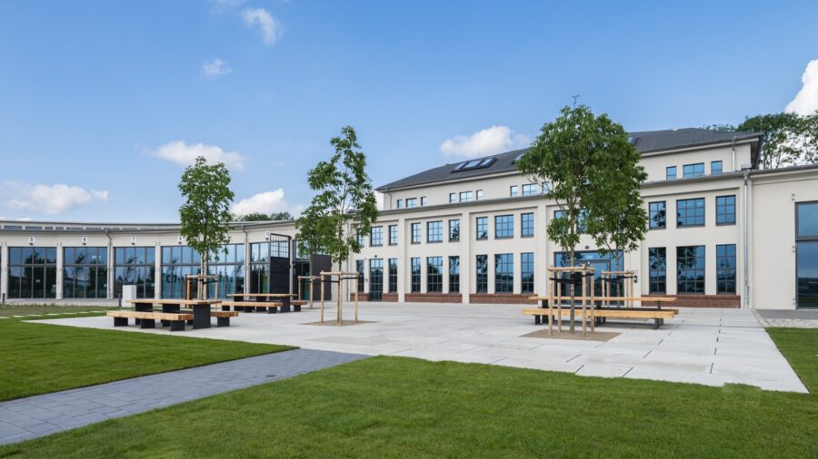 Weitwinkelaufnahme eines modern sanierten Bürogebäudes mit heller Fassade, großen blauen Fenstern, neu angelegtem Vorplatz mit Bäumen, Sitzbänken und angrenzenden Rasenflächen unter blauem Himmel.