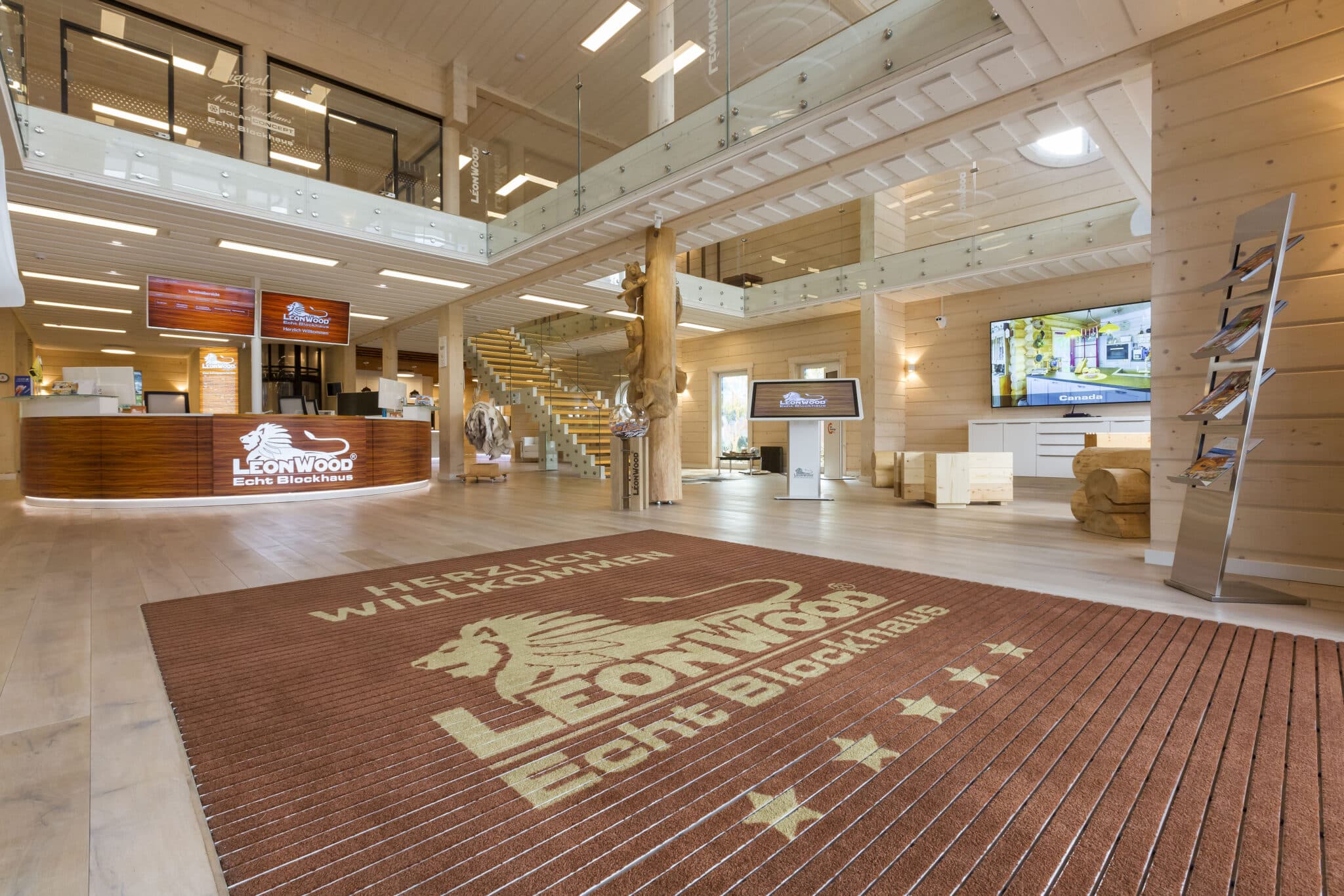 Großer, heller Empfangsbereich aus Holz bei LeonWood mit Infotheke, Treppe, Glasbalustraden und einem Teppich mit Firmenlogo.