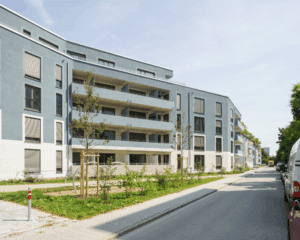 Moderne Wohnanlage mit blau-grauer Fassade, großzügigen Balkonen und begrüntem Vorgarten in städtischer Umgebung.