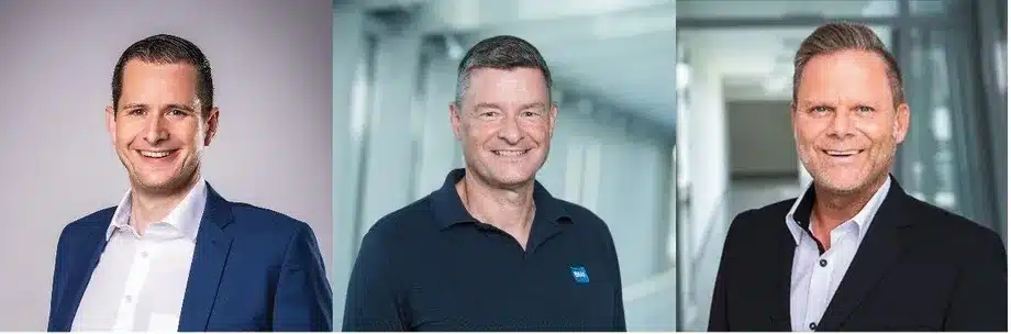 Drei Männer in Businesskleidung lächeln in die Kamera, Porträtaufnahme in professioneller Umgebung.