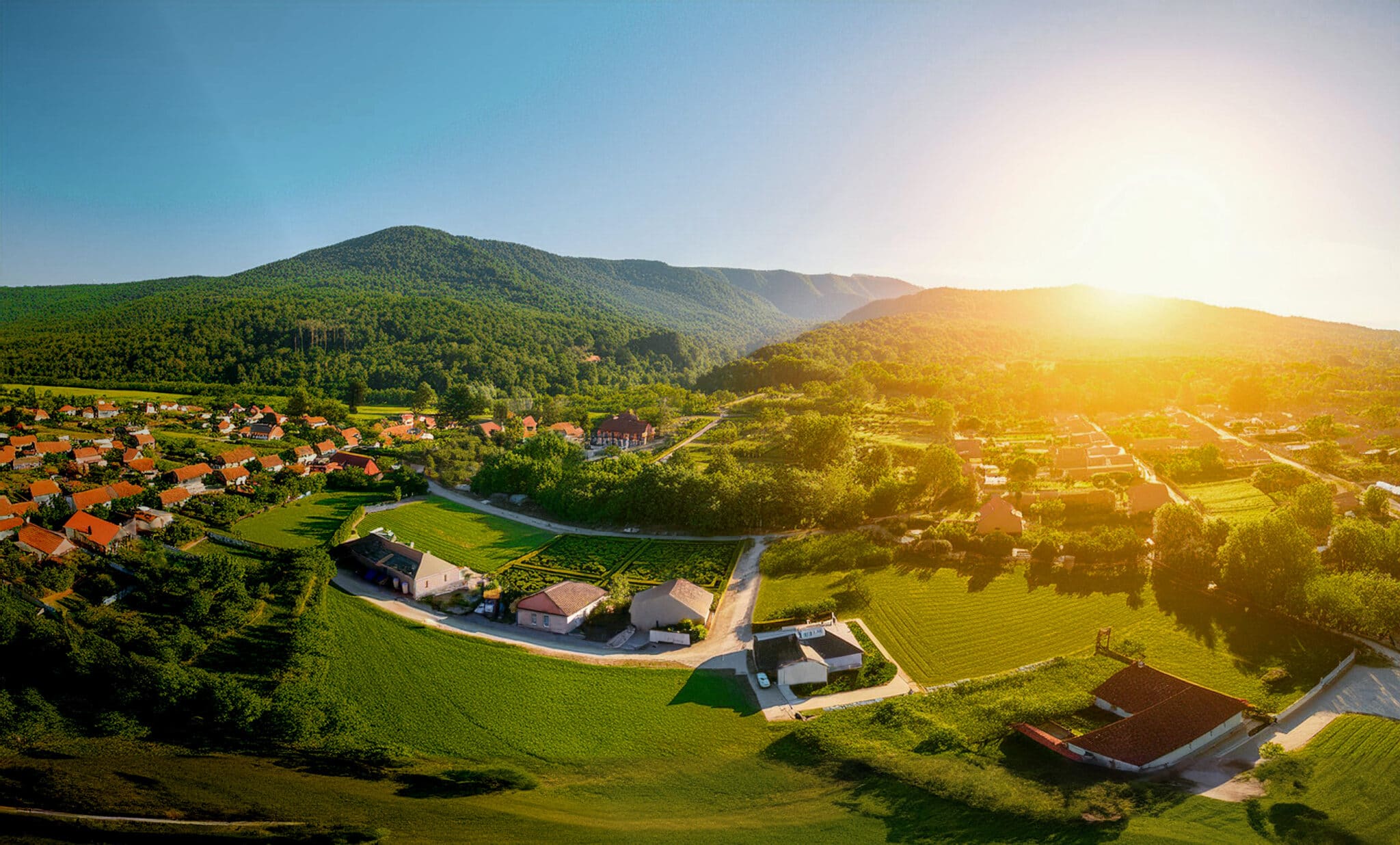 Luftaufnahme einer sonnenbeschienenen, grünen Landschaft mit Dörfern, Feldern und Wäldern in den Bergen.