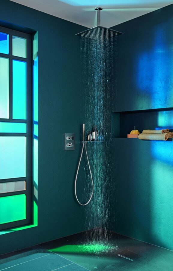 Großformatige Aufnahme einer quadratischen Regendusche, aus der ein dichter Wasserstrahl fällt. Die Dusche ist in eine dunkelgrüne Wandnische integriert, daneben farbige Glasflächen.