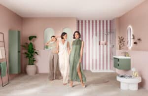 Drei stilvoll gekleidete Frauen posieren in einem modernen Badezimmer mit pastellrosa Wänden, grünem Waschtischunterschrank und stilvoller Dusche im Hintergrund.