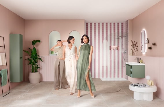 Drei stilvoll gekleidete Frauen posieren in einem modernen Badezimmer mit pastellrosa Wänden, grünem Waschtischunterschrank und stilvoller Dusche im Hintergrund.