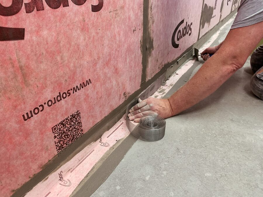 Handwerker bringt ein Schnittschutzband in frische Spachtelmasse auf einer Wand-Boden-Anschlussfuge im Bad an; Abdichtbahn von Sopro sichtbar.