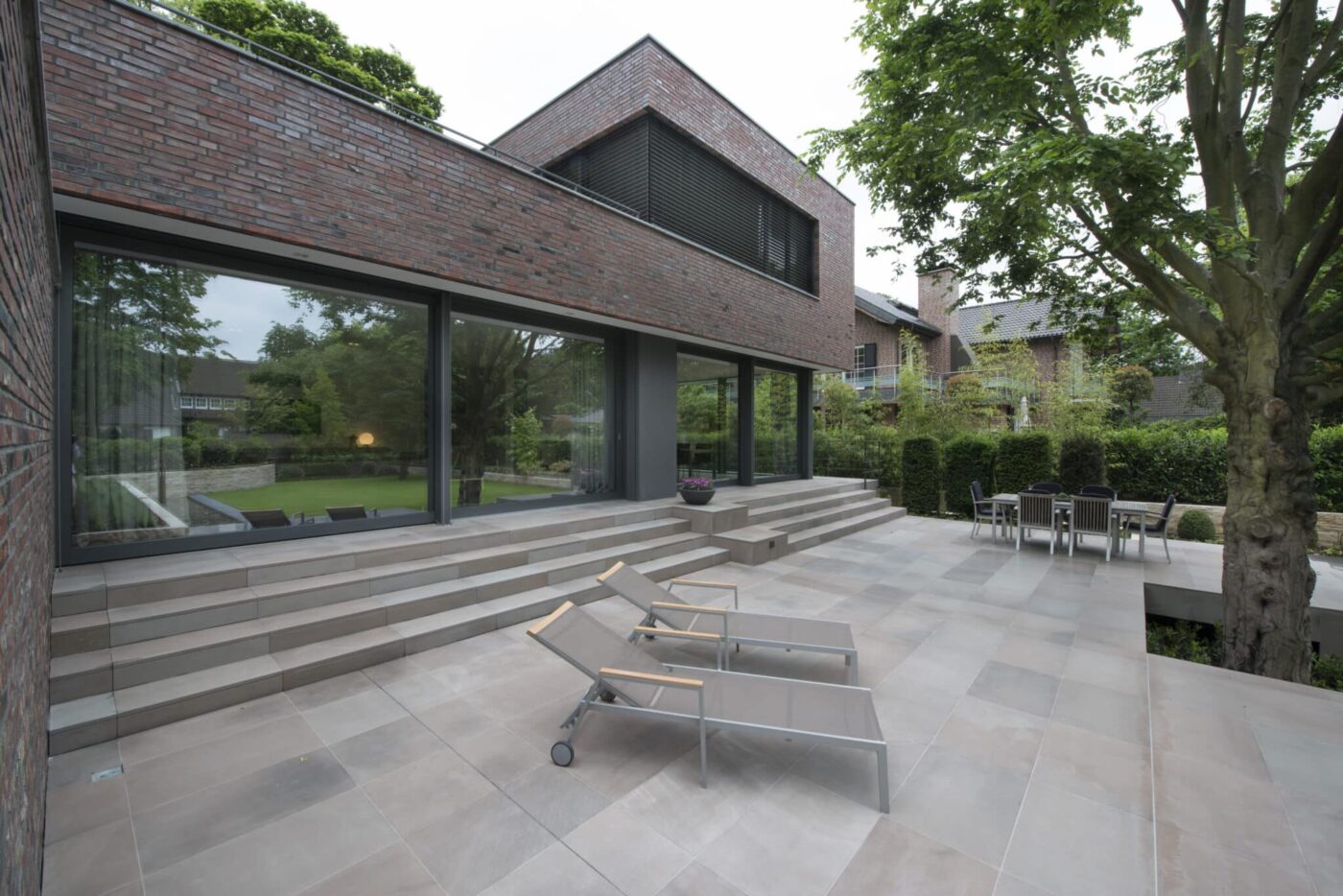 Moderne Backsteinvilla mit großer Fensterfront und weitläufiger Terrassenfläche aus großformatigen Platten, ergänzt durch Liegestühle und Gartenmöbel im grünen Außenbereich.