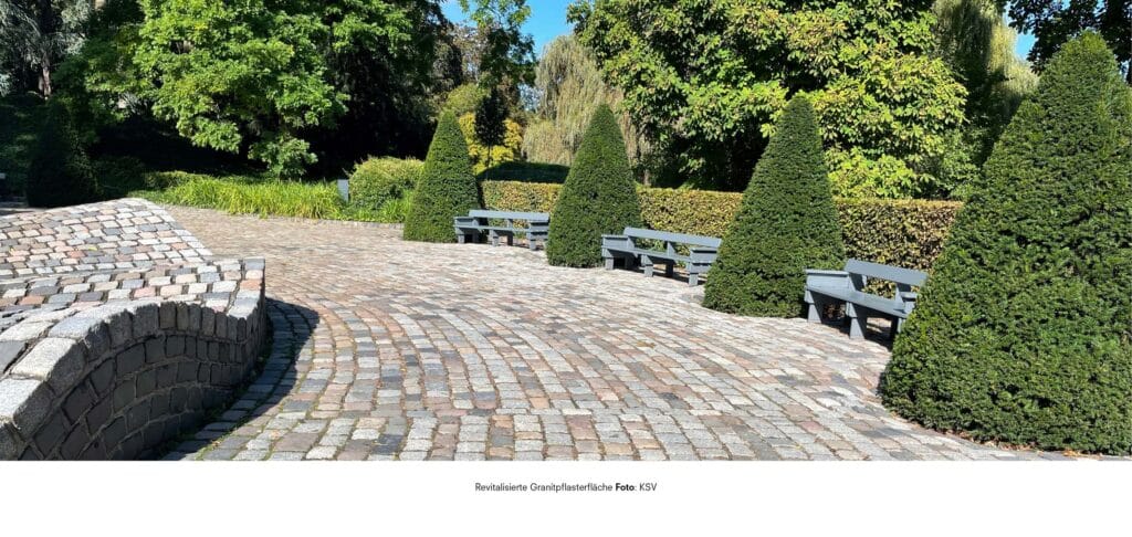 Sanierte und neu verlegte Granitpflasterfläche in einer parkähnlichen Anlage mit formgeschnittenen Gehölzen und Sitzbänken unter Bäumen.