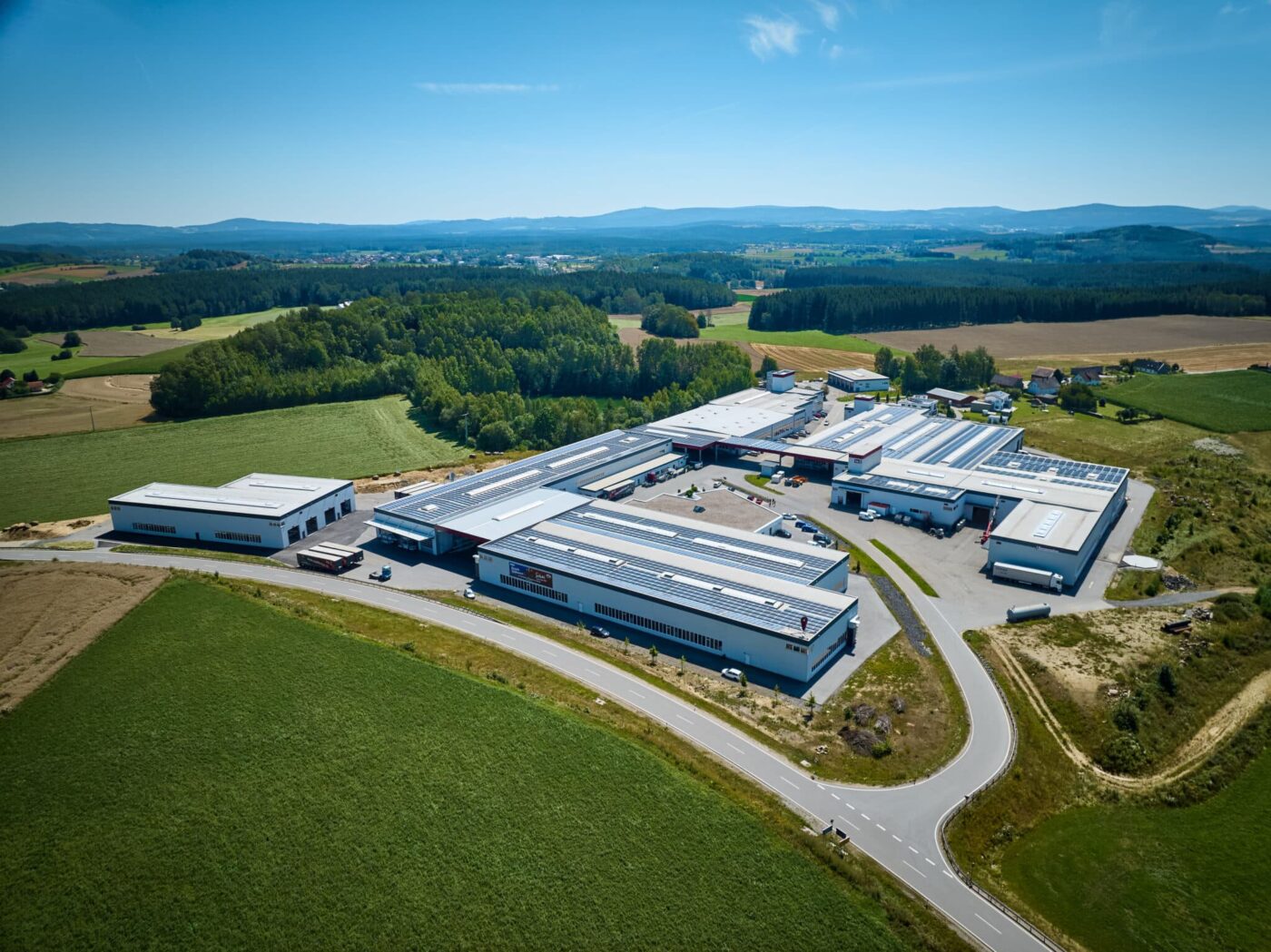 Luftaufnahme eines weitläufigen Industrie- und Produktionsstandorts, umgeben von Feldern und Wäldern; große Hallen mit mehreren Dachflächen und Logistikbereichen.