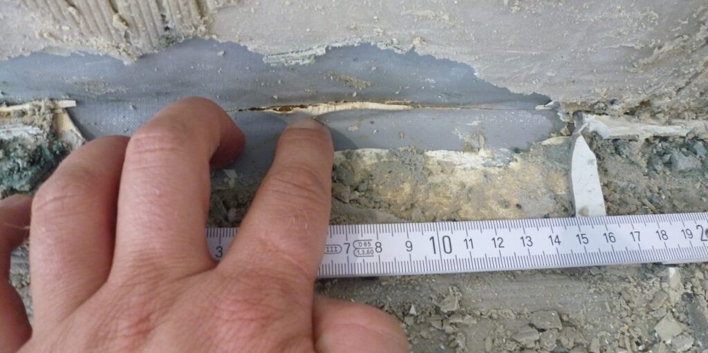 Offene und beschädigte Abdichtung unter einer Silikonfuge, freigelegt durch Entfernen des Fugenmaterials; Hand und Maßband zur Dokumentation des Schadens.
