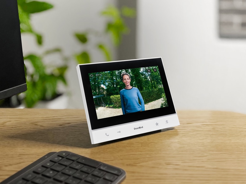 DoorBird Tisch-Innenstation auf einem Schreibtisch, zeigt das Live-Videobild einer Besucherin und steht neben einer Tastatur.