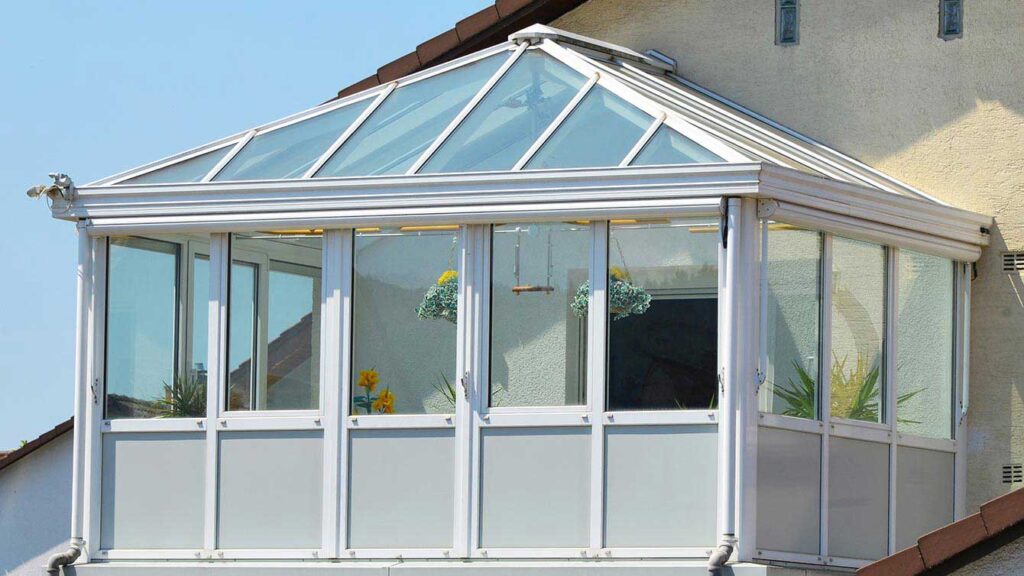 Geschlossener Wintergarten mit Glasdach und weißen Aluminiumrahmen an einem Wohnhaus, bepflanzt mit Blumen und Hängeampeln