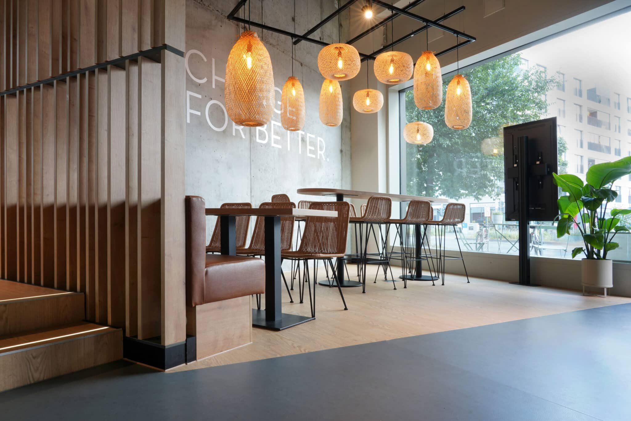 Restaurant mit Holzoptik-Boden, warmen Pendelleuchten, Naturmaterialien und modernem Sitzbereich vor großer Fensterfront.