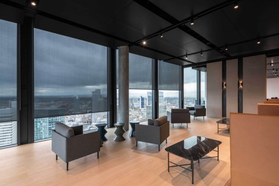 Lounge-Bereich in einem Hochhaus mit bodentiefen Fenstern, modernen Sitzmöbeln und textilen Sonnenschutzrollos.