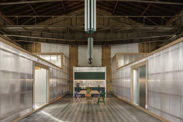 Zwei lichtdurchlässige Haus-im-Haus-Volumen in einer Industriehalle mit zentraler Küche und Essbereich.