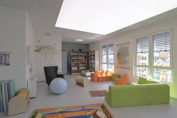 Helle offene Kommunikationsflächen in einer Grundschule mit bunten Sitzmöbeln, Bücherregalen und viel Tageslicht.